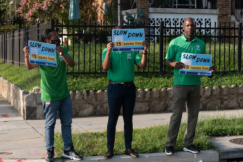 Volunteers stand on a corner holding up signs asking drivers to slow down to help students arrive at school safely. (Shevry Lassiter/The Washington Informer)