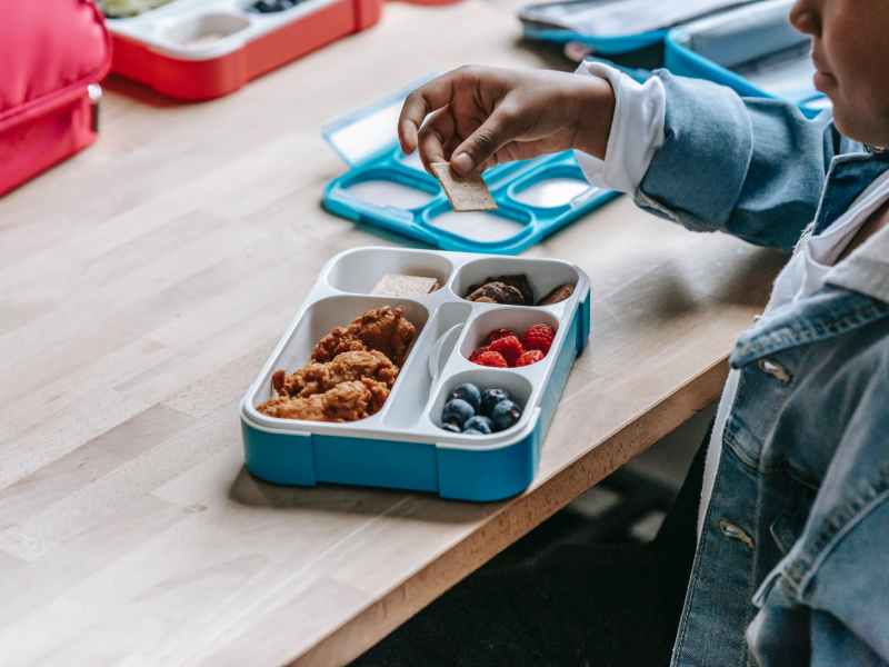 crop black schoolchild with biscuit near lunch box