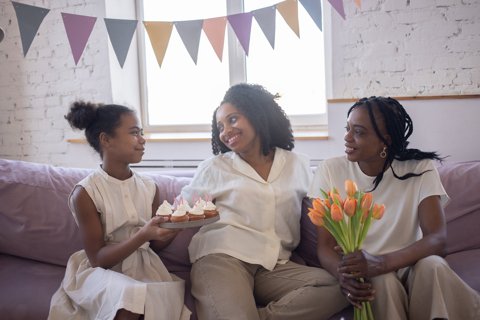 a young girl in white dress holding a plate with cupcakes while looking at the women sitting on the couch