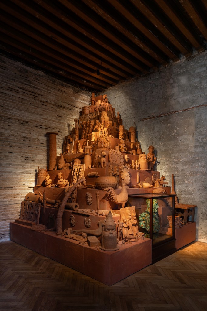 The artwork is “Monument to the Restitution of the Mind and Soul, 2023” by Yinka Shonibare, a dynamic terracotta sculpture that intricately weaves figures and architectural forms into a city-like structure, exhibited against the raw textures of a rustic space.(Courtesy of the Museum of West African Art)