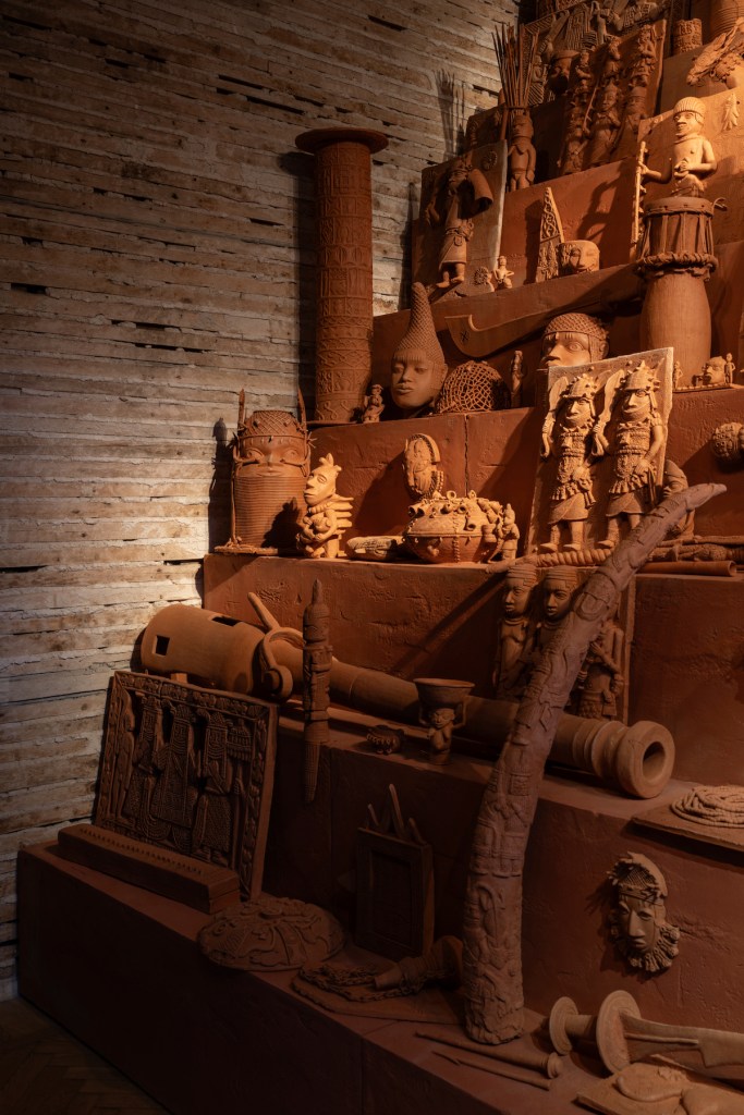 “Monument to the Restitution of the Mind and Soul, 2023” by Yinka Shonibare is an evocative terracotta sculpture displayed here. This detailed piece features an array of cultural and historical elements, constructing a narrative that climbs upwards in a layered, city-like formation, all captured under the warm glow of exhibition lighting.(Courtesy of the Museum of West African Art)