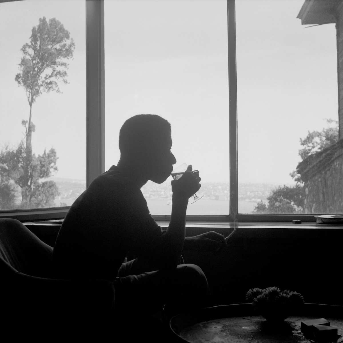 James Baldwin, Istanbul, c. 1965. A reflective moment captured by Sedat Pakay in a gelatin silver print, showing Baldwin’s silhouette seated by a large window, sipping a drink and smoking, with a serene landscape view in the background.(Courtesy of Sedat Pakay)